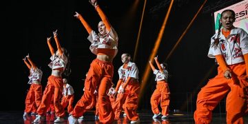 „M-BEATS“ šokių studijos vaikų grupė oranžinėmis kelnėmis ir šviesiais margintais marškinėliais scenoje atlieka bendrą choreografiją